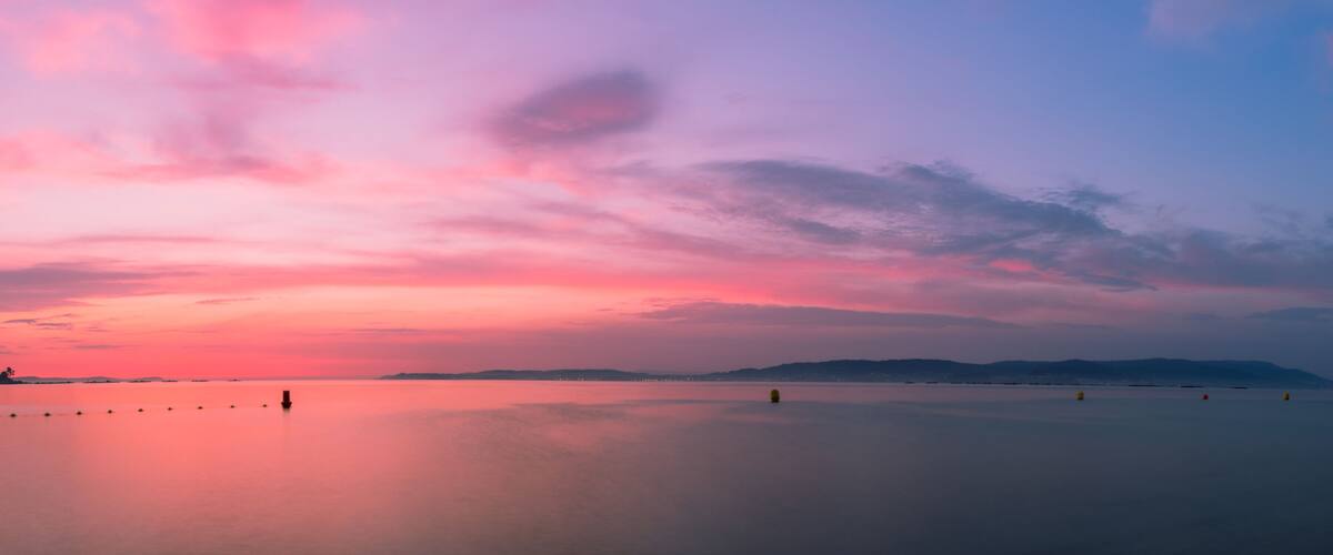 A calm lake at sunset with a pink and purple sky, distant mountains, and floating buoys.