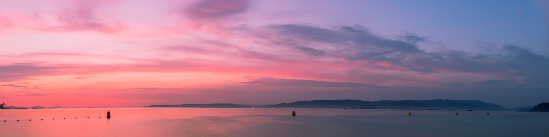 A calm lake at sunset with a pink and purple sky, distant mountains, and floating buoys.