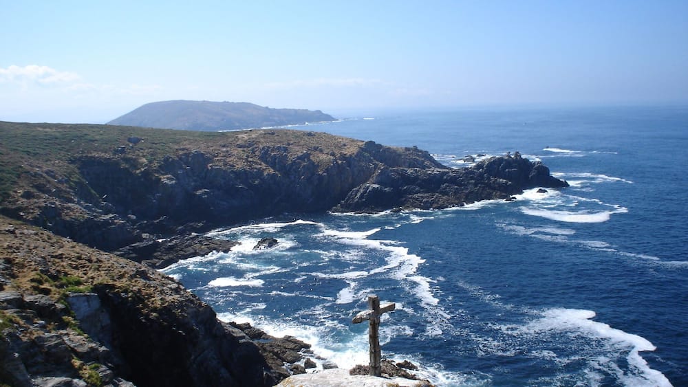 A coastal landscape with rocky cliffs, waves crashing against the shore, and a cross on a rock.