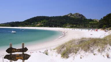 A beach with a sign indicating Praia de Rodas, surrounded by clear turquoise waters, white sand, and a forested hill in the background.
