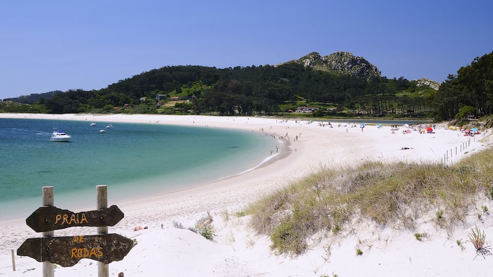 A beach with a sign indicating Praia de Rodas, surrounded by clear turquoise waters, white sand, and a forested hill in the background.