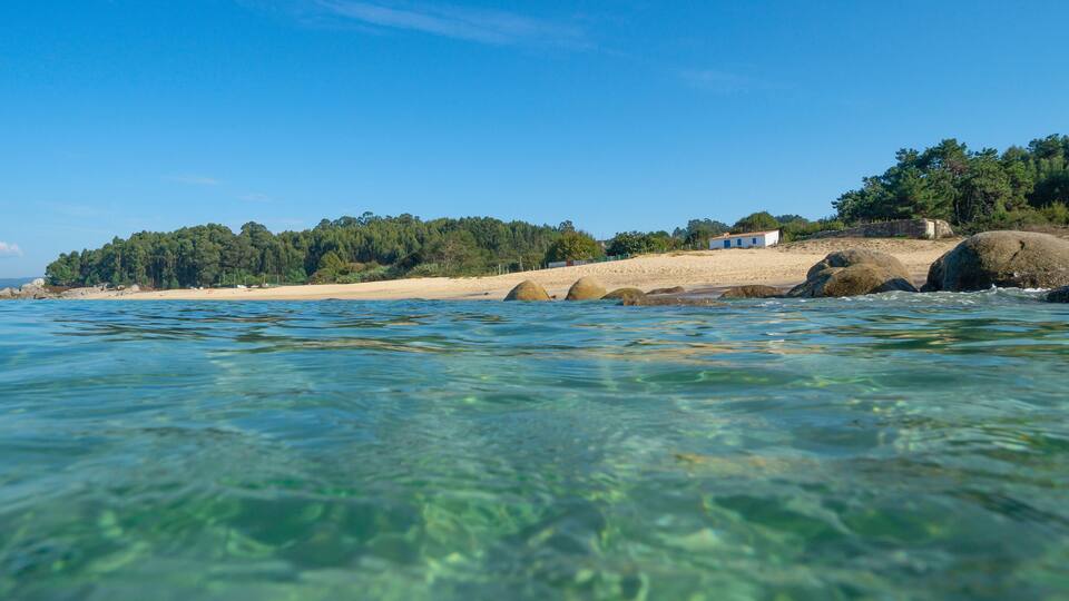 A clear coastal view with a sandy beach, large rocks, and a small building on a hill.