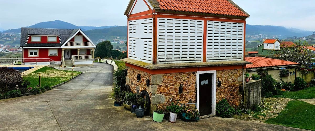 A stone church with a red roof and white shutters, surrounded by houses and a mountainous landscape.