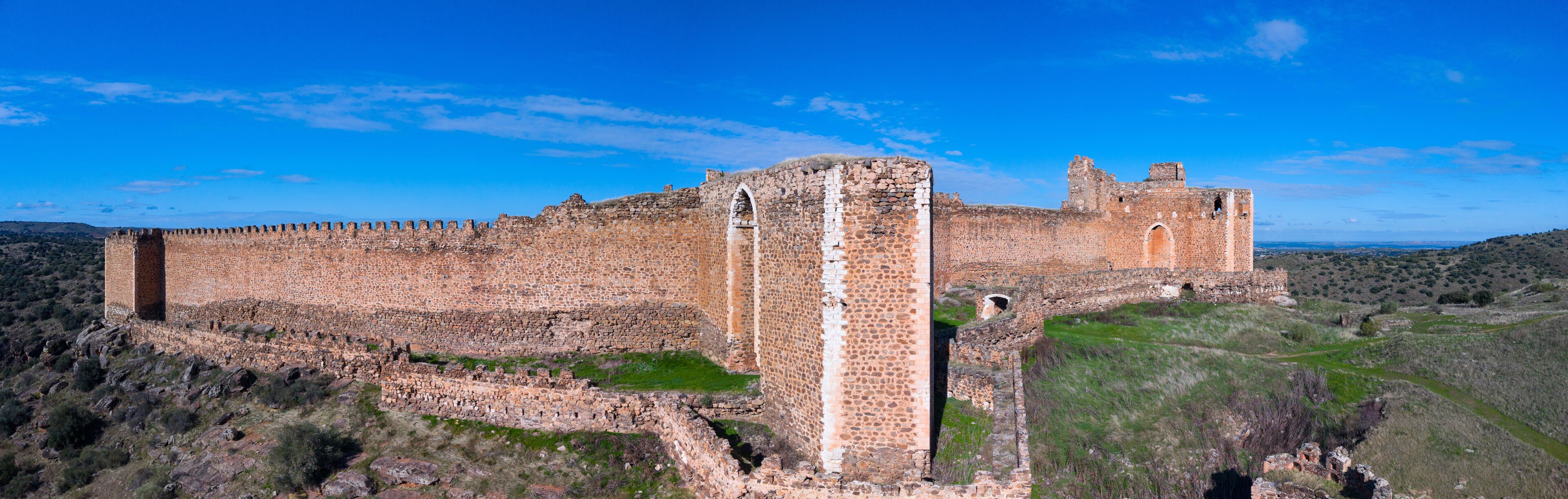 A large, ancient stone fortress with a prominent central tower and a surrounding wall, set against a clear blue sky and hilly landscape.