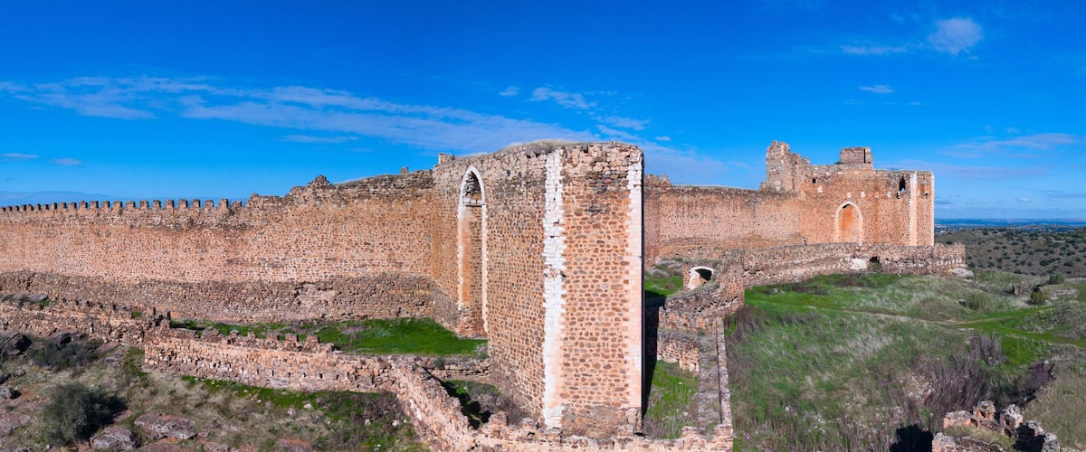 A large, ancient stone fortress with a prominent central tower and a surrounding wall, set against a clear blue sky and hilly landscape.