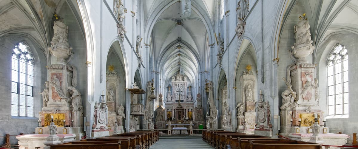 A cathedral interior with high vaulted ceilings, rows of pews, and ornate statues.