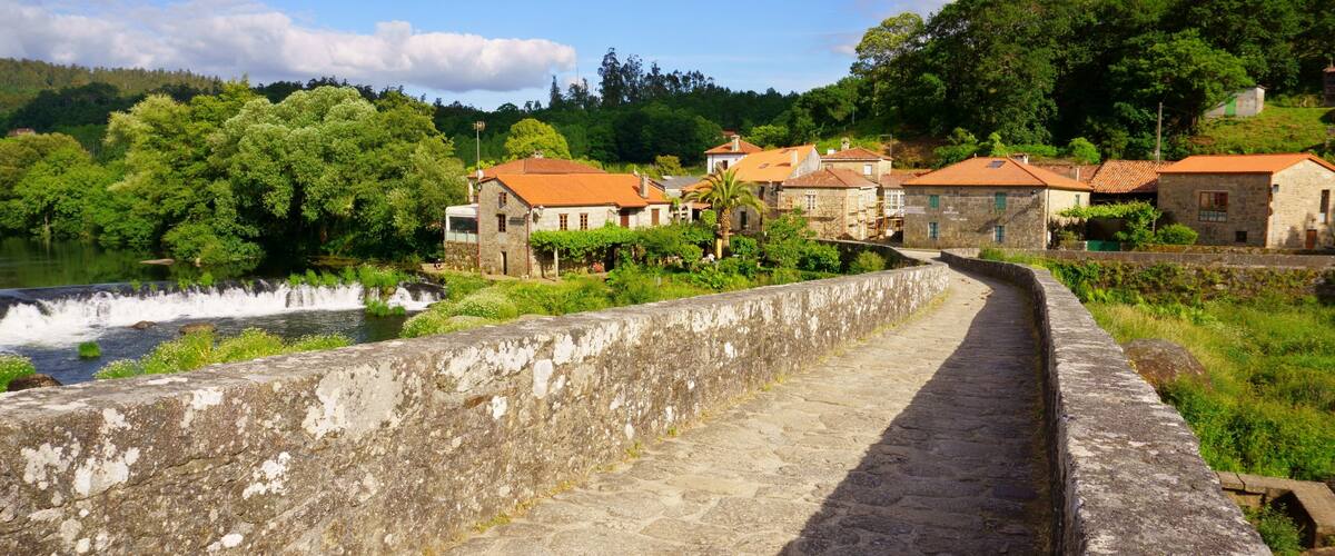 A stone pathway beside a river, with traditional buildings and lush greenery in the background.