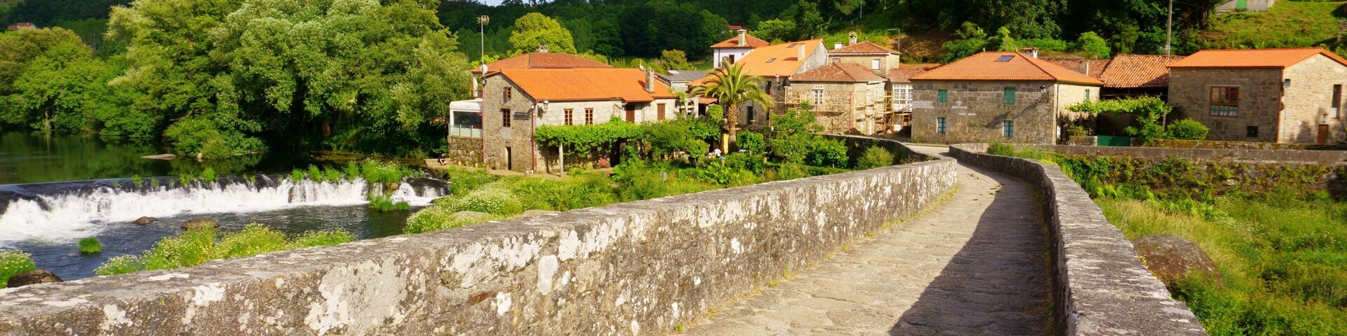 A stone pathway beside a river, with traditional buildings and lush greenery in the background.