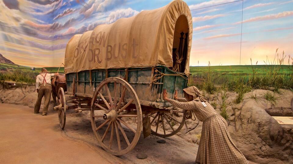 A covered wagon with 'OREGON OR BUST' written on it, pulled by a horse, with a person in a checkered dress standing beside it.