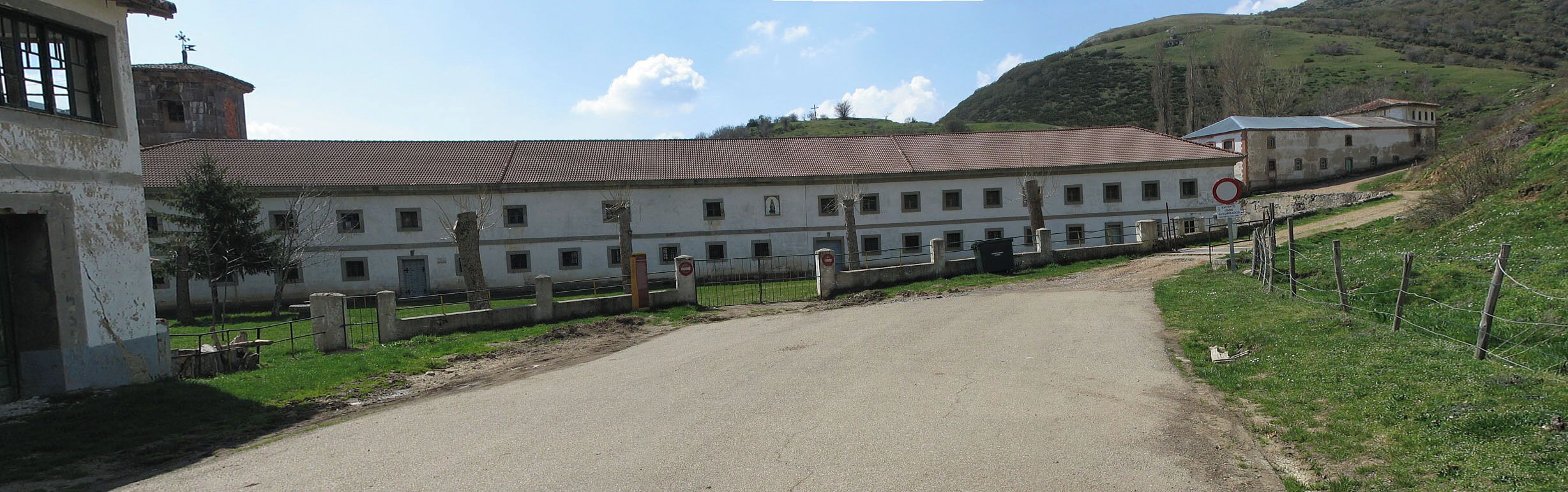 A large white building with a red-tiled roof, surrounded by a fence and a dirt path leading to a road.