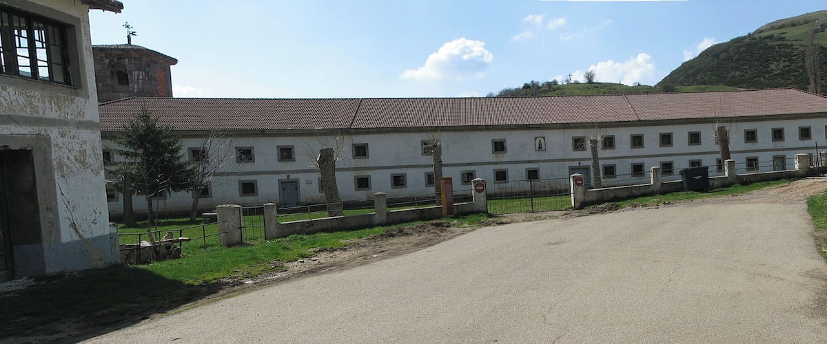 A large white building with a red-tiled roof, surrounded by a fence and a dirt path leading to a road.