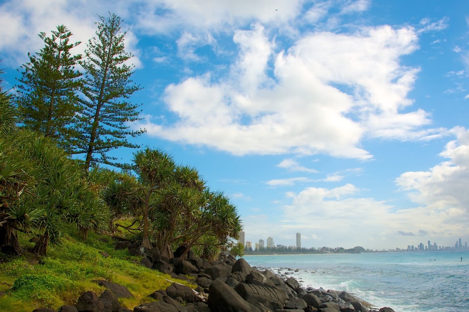 A coastal landscape with rocky shores, dense green foliage, and a clear blue sky with scattered clouds.
