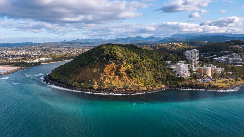 An aerial view of a coastal city with a prominent hill, a beach, and a clear blue sea.