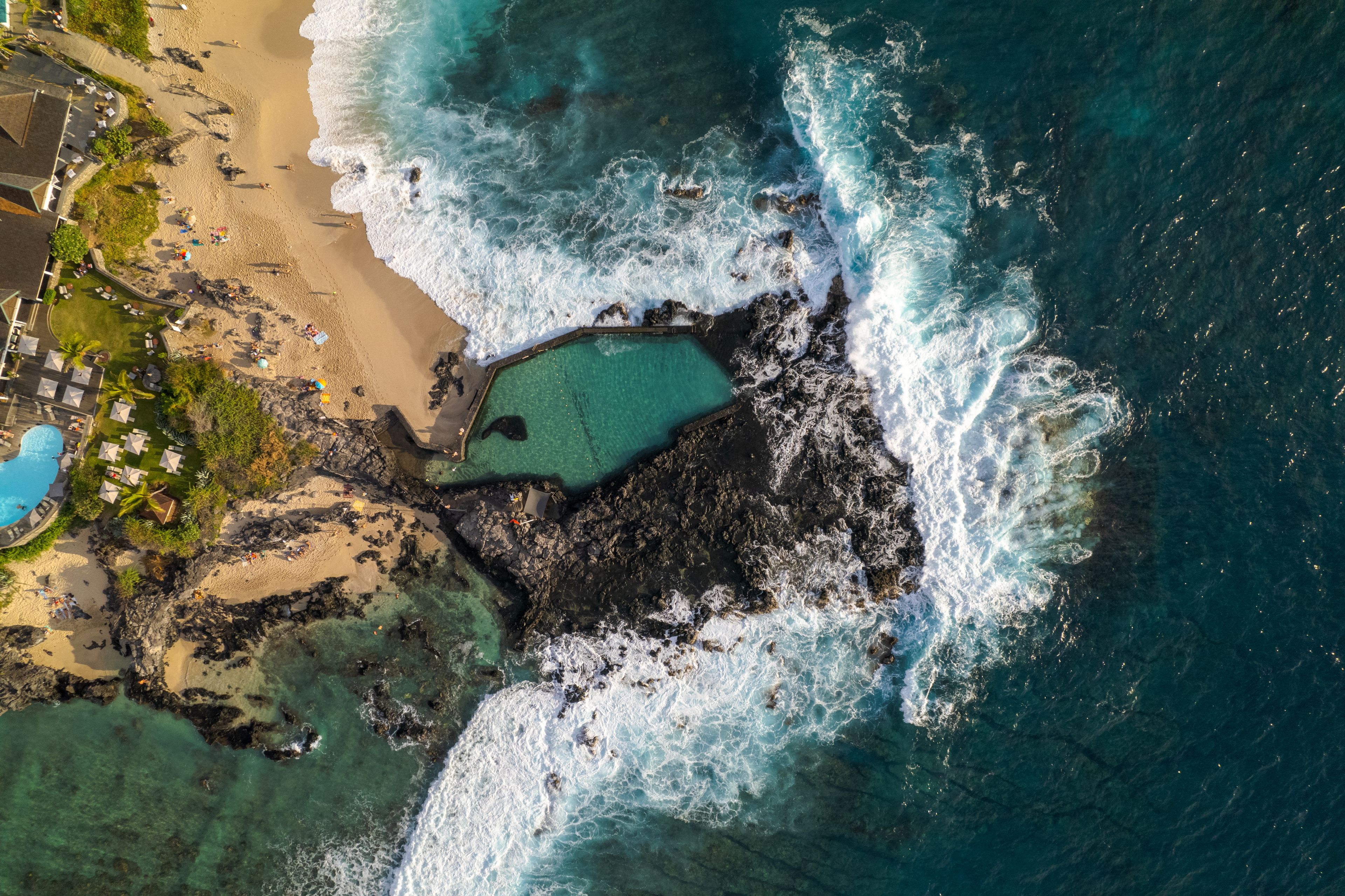 Una zona costera con costa rocosa, una piscina azul cristalina y una playa de arena con sombrillas.