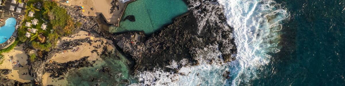 A coastal area with a rocky shoreline, a clear blue pool, and a sandy beach with umbrellas.