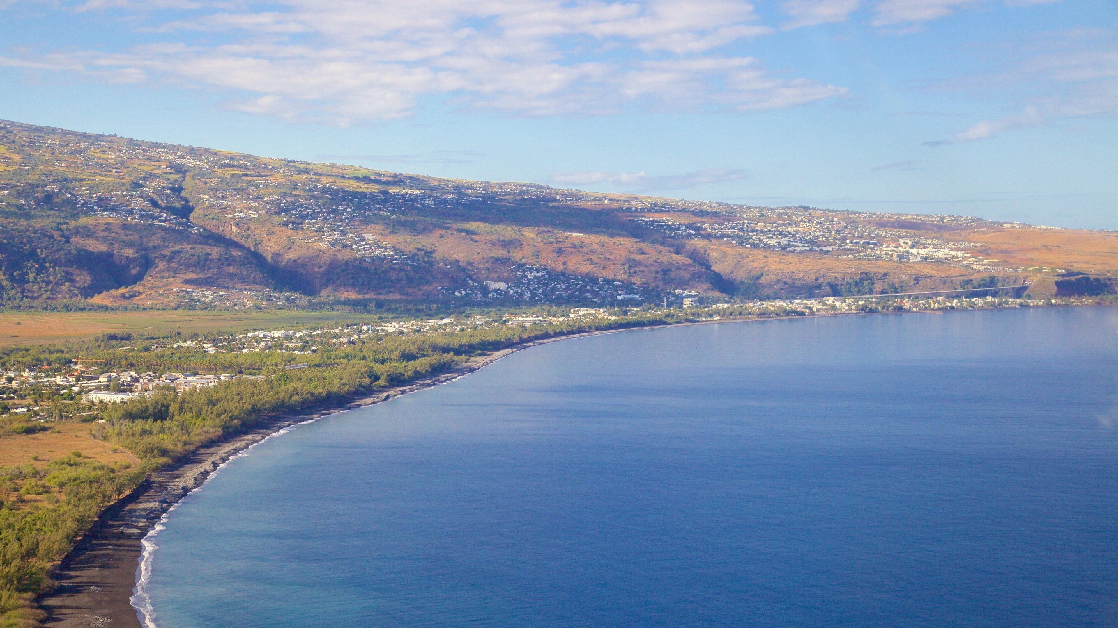 A coastal town with a curved shoreline, a bridge, and a hilly landscape in the background.