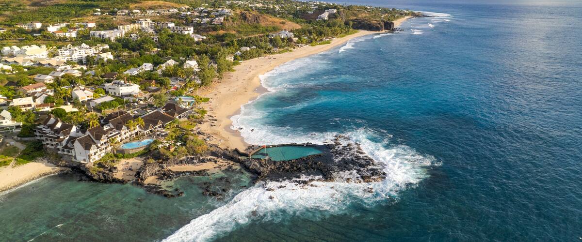 Un pueblo costero con playa, océano y un promontorio rocoso con piscina.