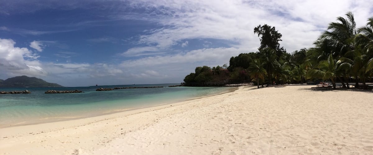 A beach with white sand, clear turquoise water, palm trees, and rocky outcrops in the distance.