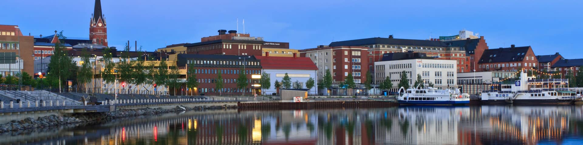 A cityscape with a prominent church steeple, a waterfront, and various buildings reflecting on the calm water.