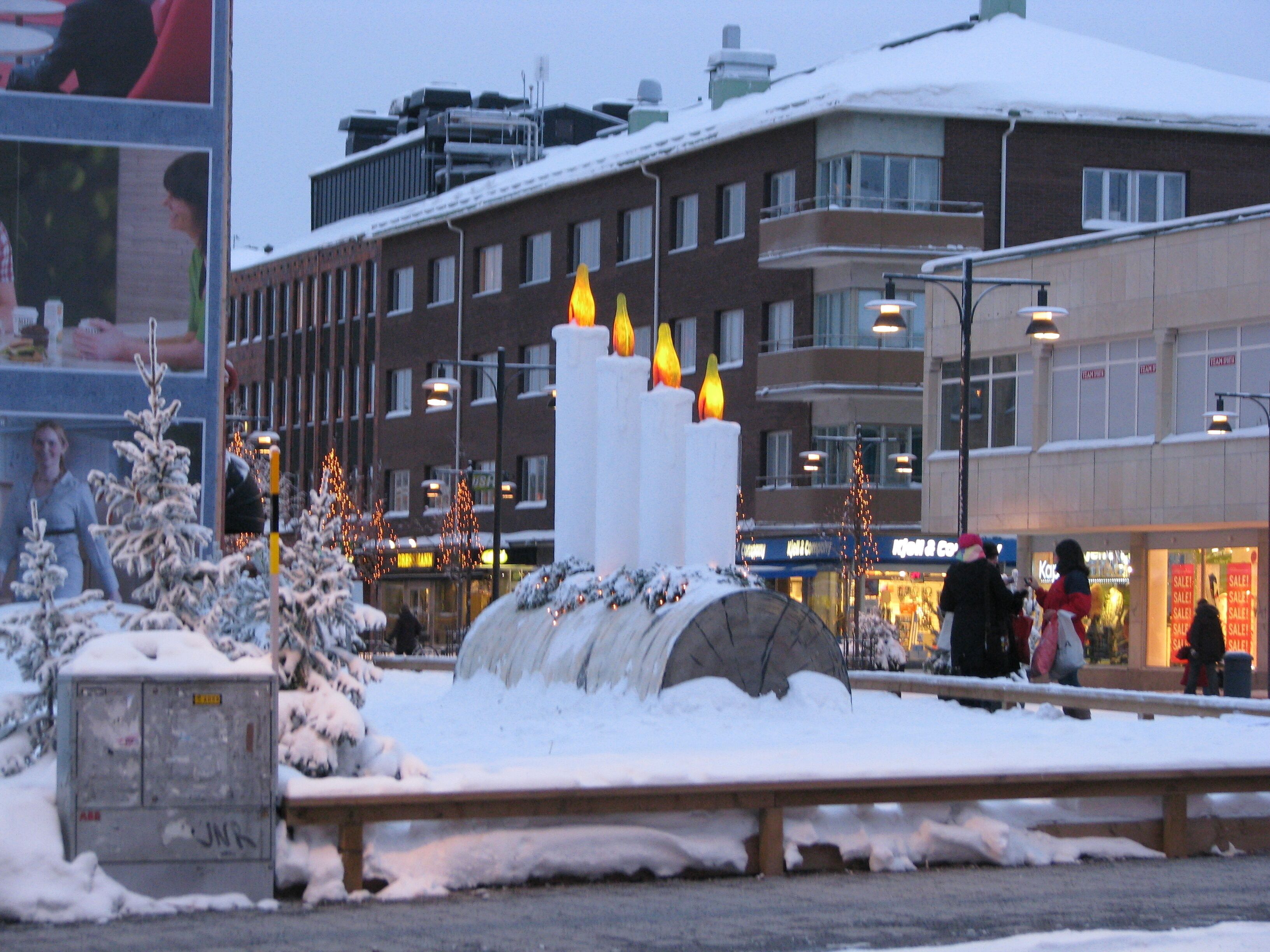 A snowy urban square with a large illuminated sculpture, buildings, and a shopping area in the background.