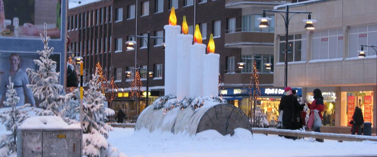 A snowy urban square with a large illuminated sculpture, buildings, and a shopping area in the background.