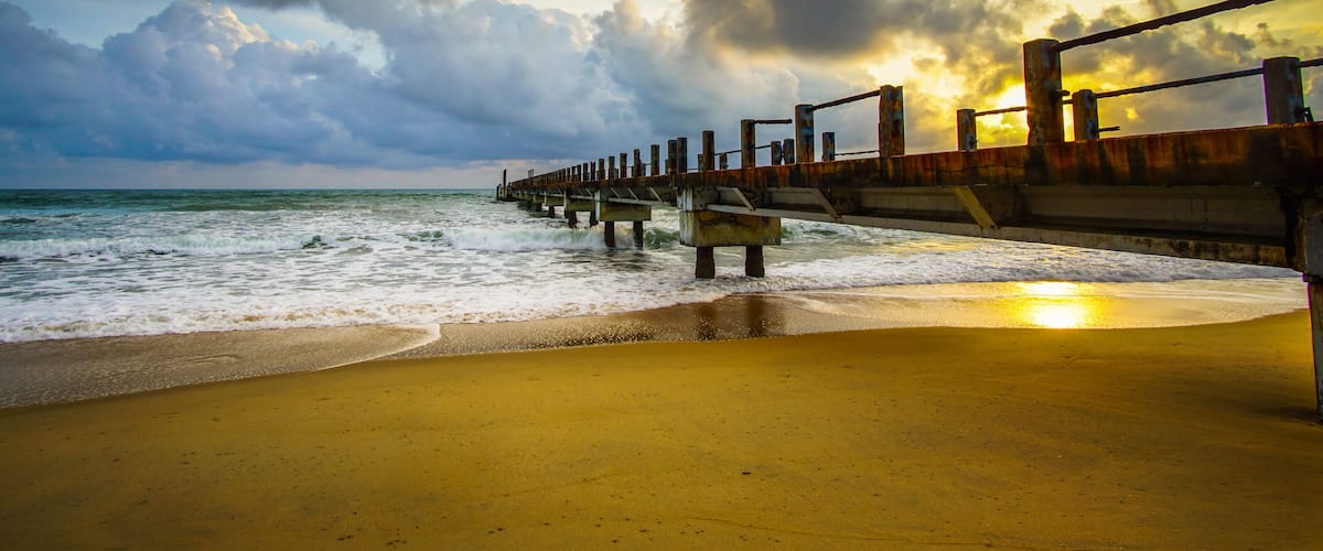 A beach with a wooden pier extending into the ocean, waves crashing against the shore, and a sunset in the background.