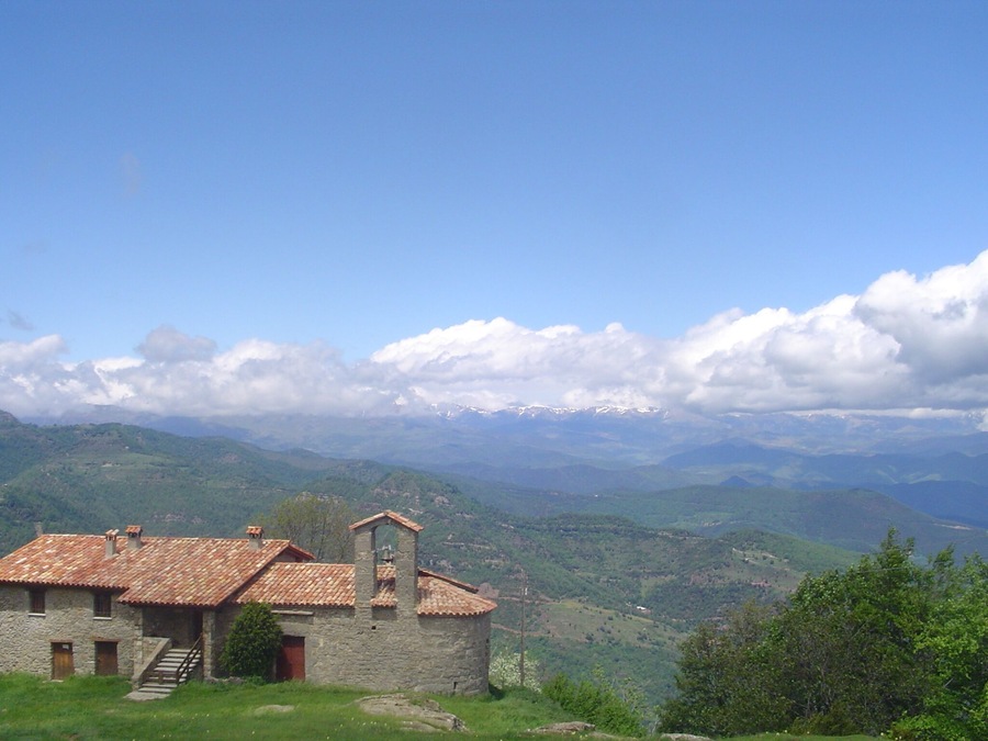 A stone house with a tiled roof, surrounded by lush green hills and distant snow-capped mountains under a clear blue sky.