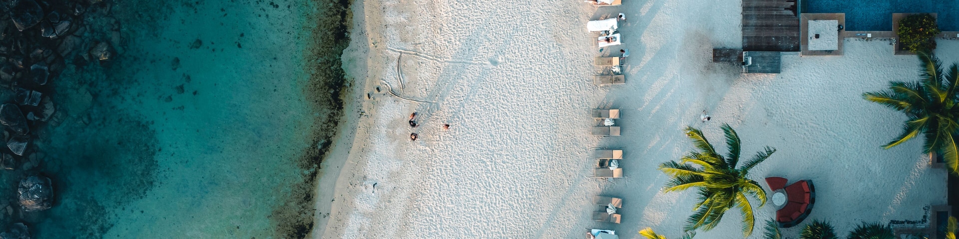 Vista aérea de una playa con agua turquesa cristalina, arena blanca y palmeras. Se observan sillas de playa y sombrillas a lo largo de la orilla, y un edificio con piscina a la derecha.