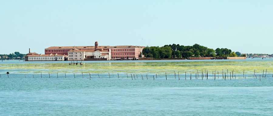 Un edificio storico con tetto rosso e pareti bianche, affacciato su una superficie d'acqua tranquilla con pontili galleggianti.