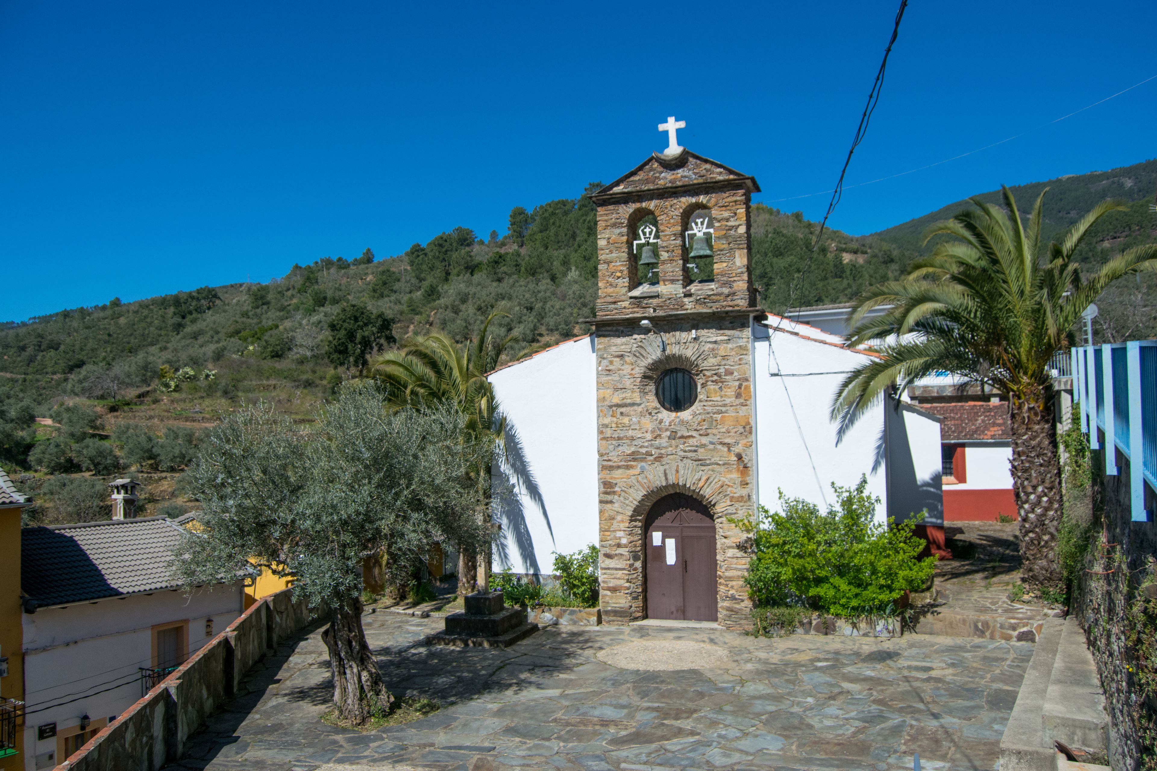 A church with a stone bell tower and a white facade, surrounded by palm trees and a hillside.