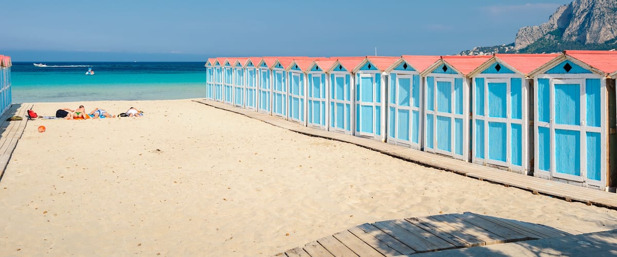 A beach with a wooden walkway lined with blue and white beach huts, a clear blue sea, and a rocky cliff in the background.