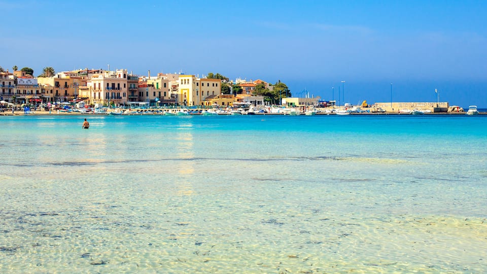 A coastal town with clear turquoise waters, a marina with boats, and colorful buildings along the shore.