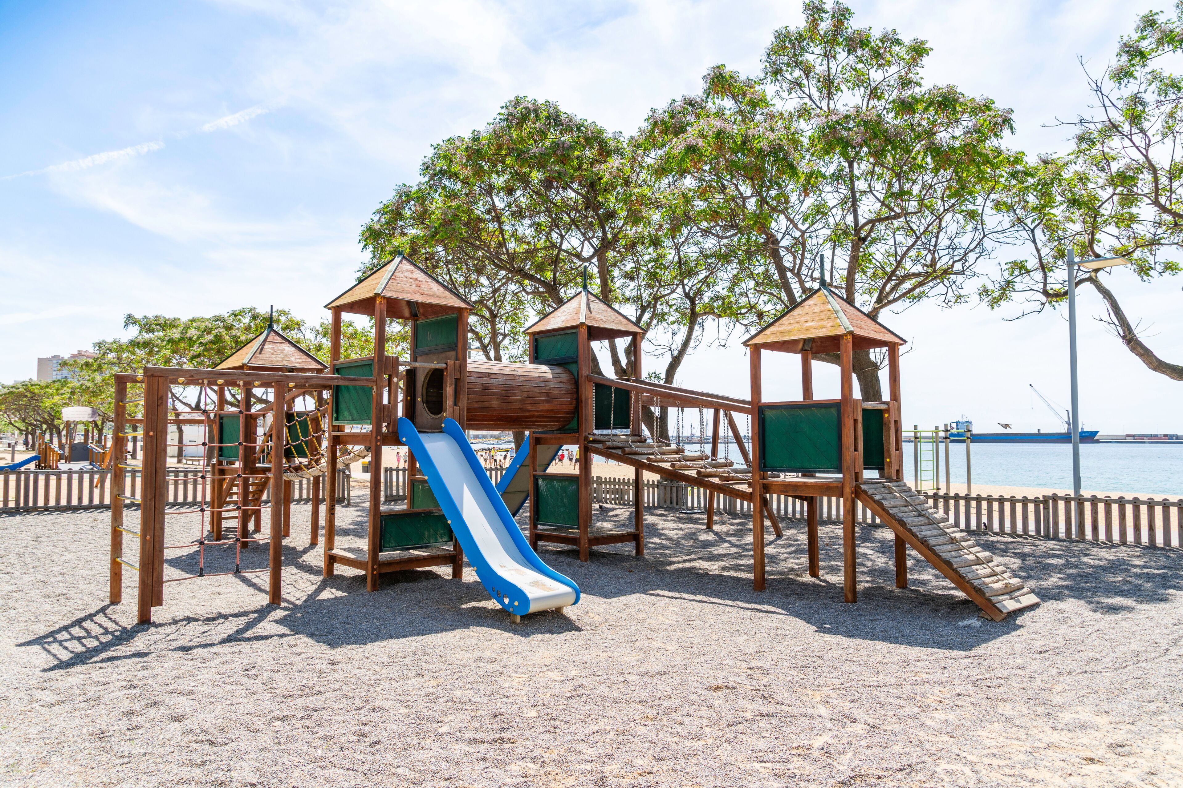 A wooden playground with a slide and climbing structures on a sandy beach with trees and a body of water in the background.