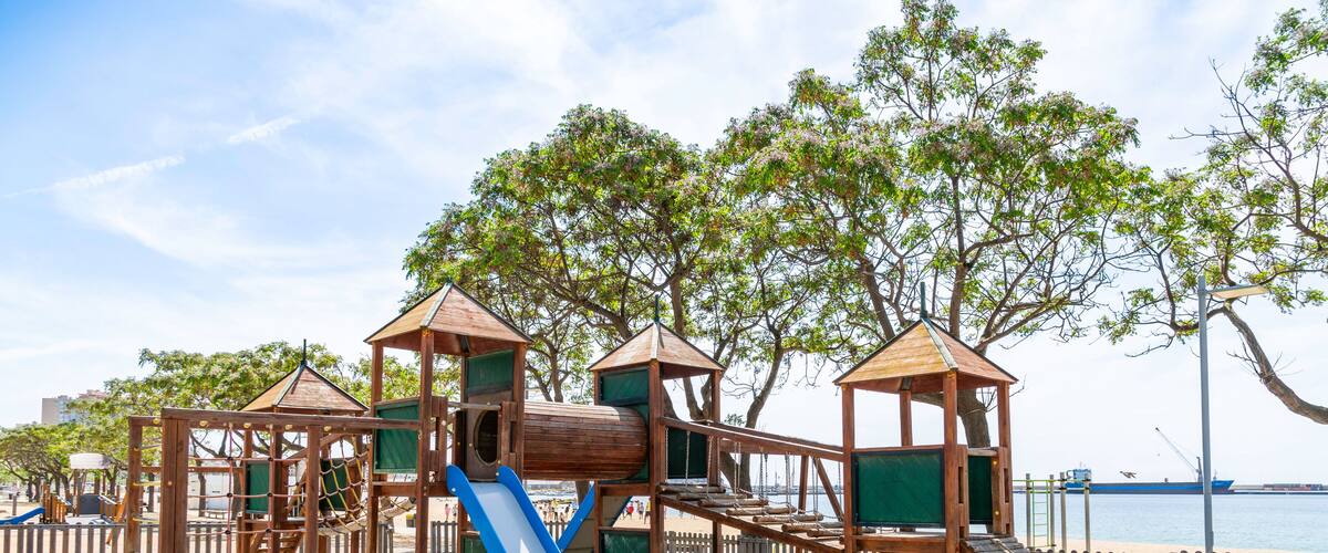 A wooden playground with a slide and climbing structures on a sandy beach with trees and a body of water in the background.