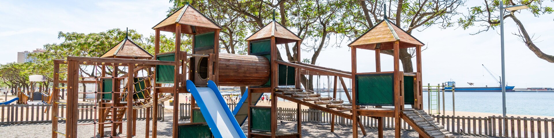 A wooden playground with a slide and climbing structures on a sandy beach with trees and a body of water in the background.