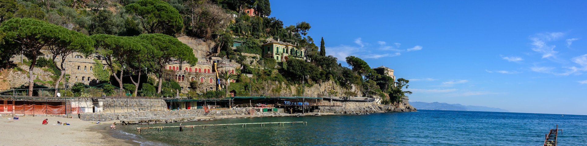 Un pueblo costero con edificios en una ladera, una playa y un muelle que se adentra en el mar.