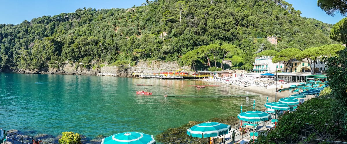 A coastal area with a beach, turquoise umbrellas, boats, and a forested hill.