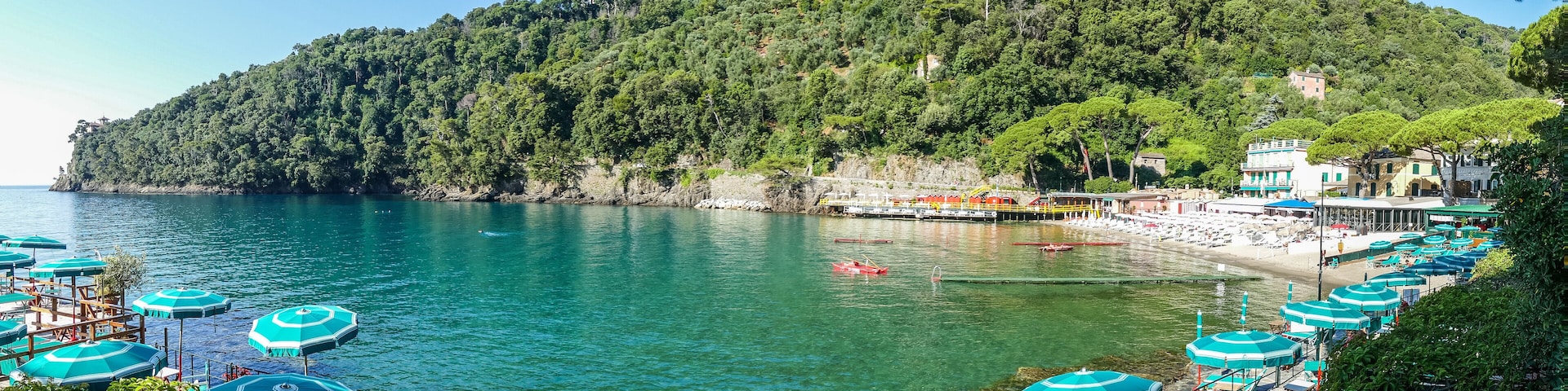 A coastal area with a beach, turquoise umbrellas, boats, and a forested hill.