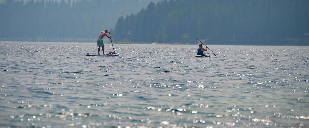 Two people paddleboarding on a calm lake with a forested mountain range in the background.
