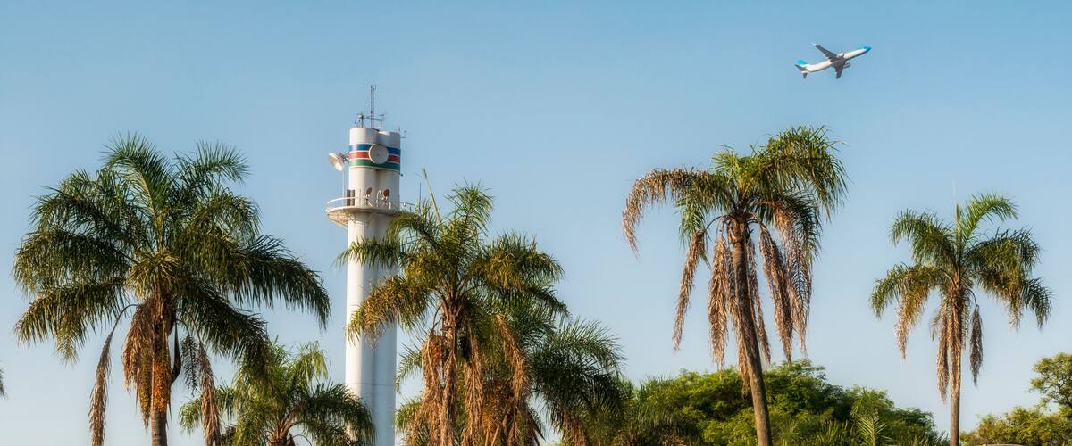 A lighthouse surrounded by palm trees and a clear blue sky with an airplane flying overhead.