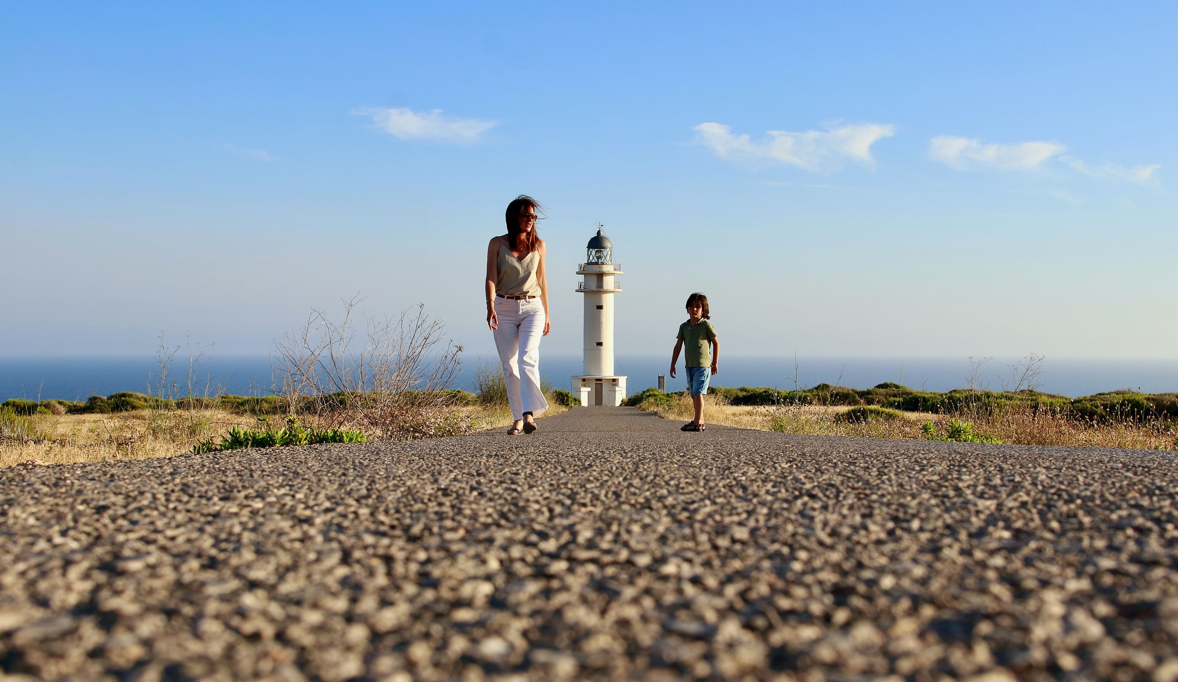 A coastal road with a lighthouse in the distance, surrounded by sparse vegetation and a clear sky.