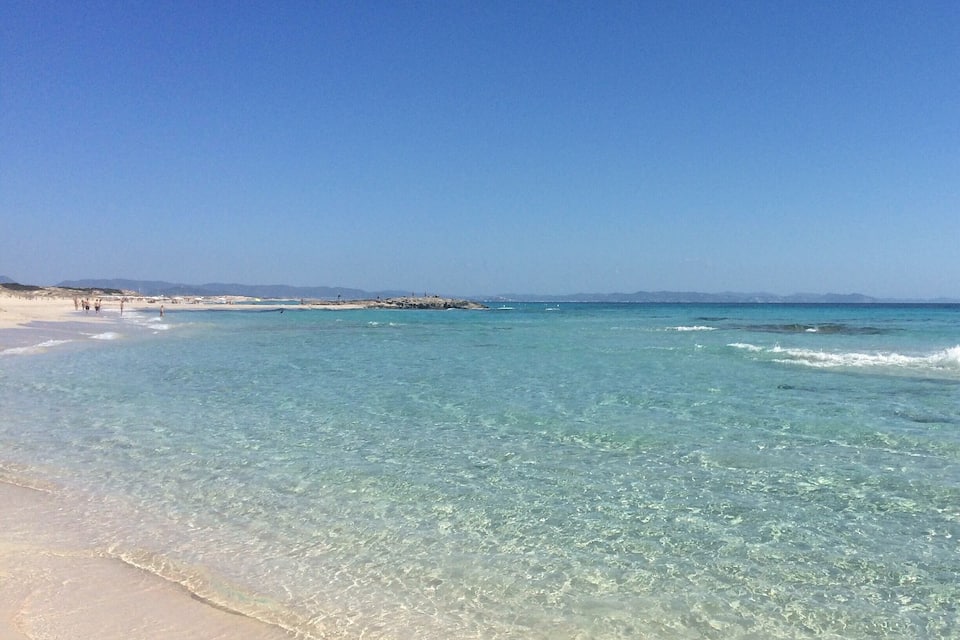 A beach with clear turquoise water, a sandy shore, and a rocky breakwater in the distance.