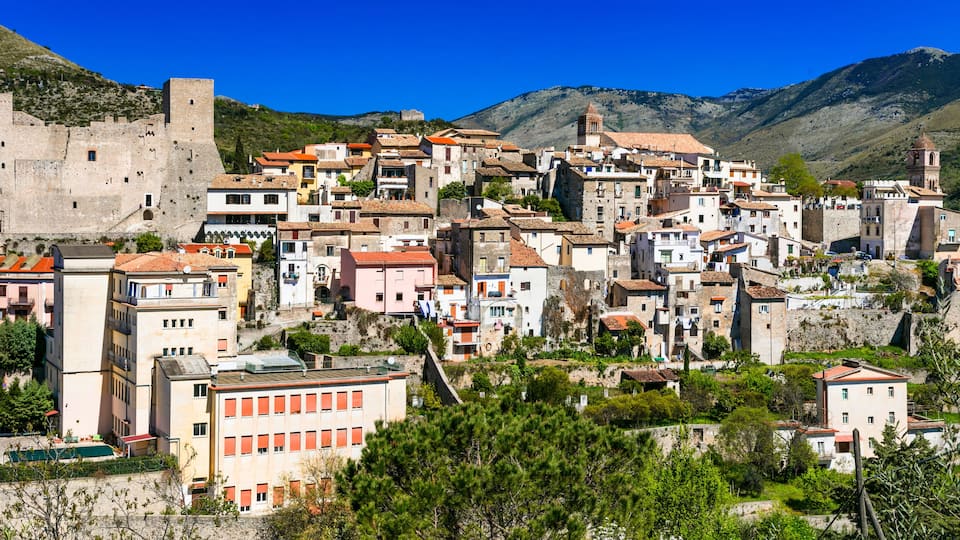 A historic town with a prominent castle, terracotta-roofed buildings, and lush green hills in the background.