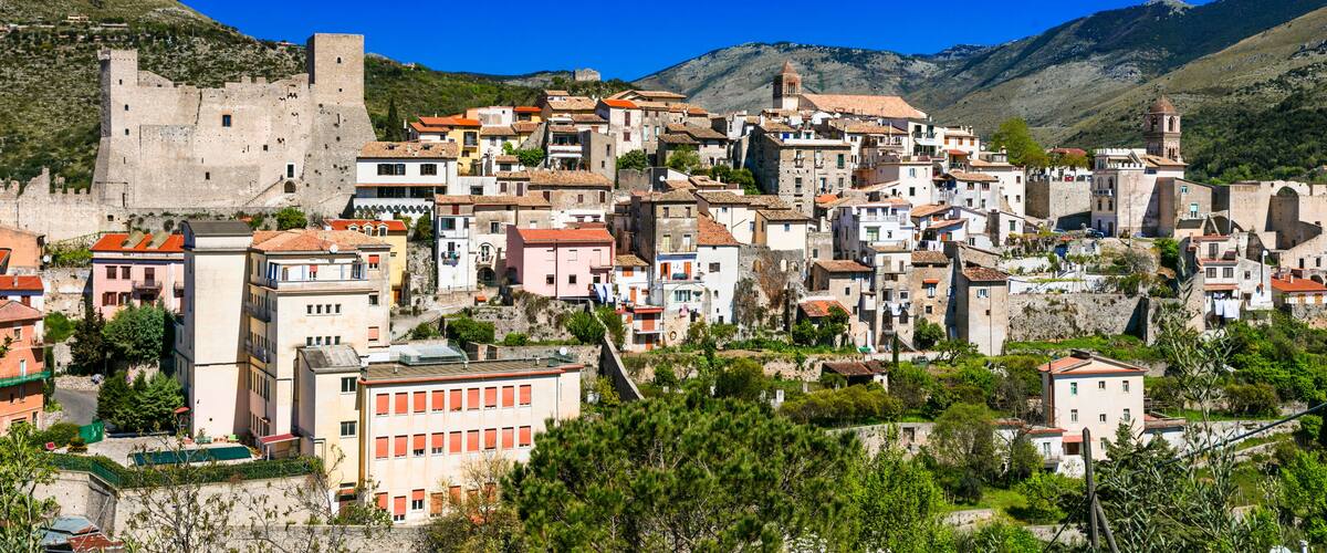 A historic town with a prominent castle, terracotta-roofed buildings, and lush green hills in the background.