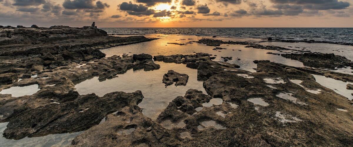 A rocky shoreline at sunset with the sun reflecting off the water.