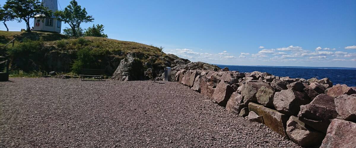 A rocky shoreline with a lighthouse, a bench, and a clear blue sky.