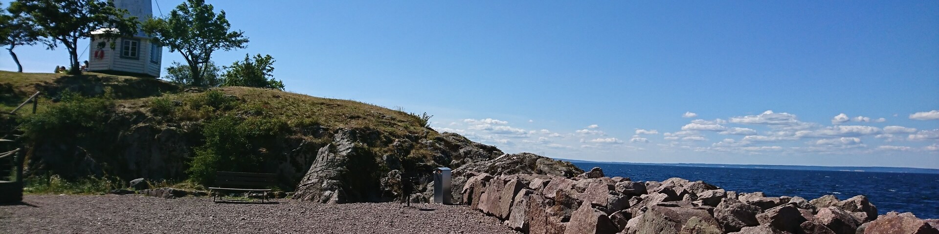 A rocky shoreline with a lighthouse, a bench, and a clear blue sky.