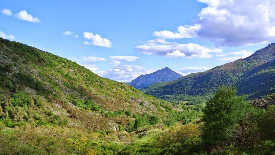 A mountainous landscape with a valley, dense greenery, and a prominent mountain peak under a partly cloudy sky.