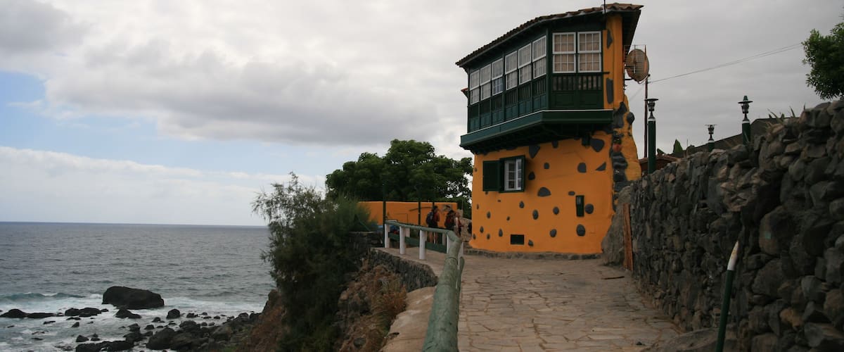 A coastal walkway with a stone wall, a yellow building with green accents, and the ocean in the background.