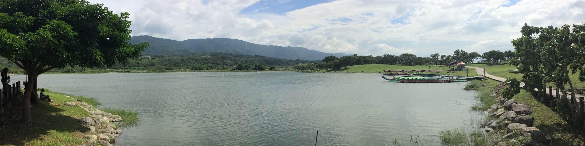 Um lago tranquilo com um píer de madeira, um barco e uma cordilheira ao longe sob um céu parcialmente nublado.